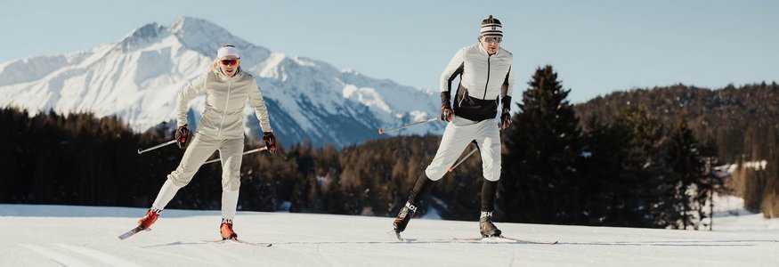 Two cross-country skiers in snow with snowy mountains and clear sky background
