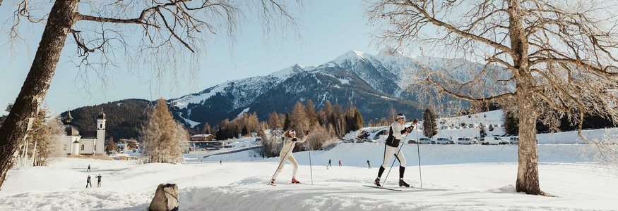 Two cross-country skiers on snow with mountains and clear sky