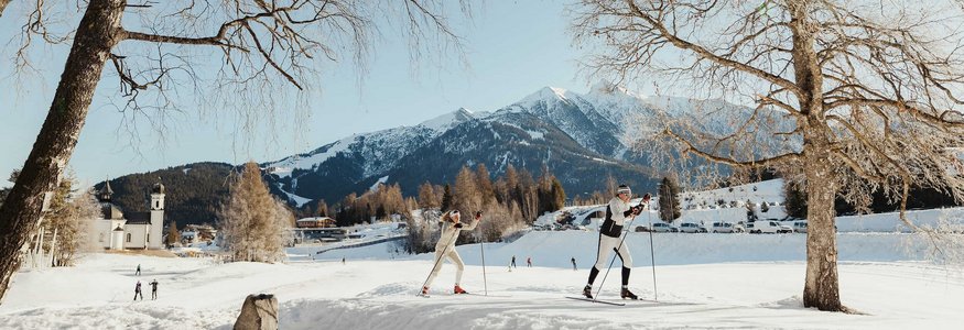 Two cross-country skiers on snow with mountains and clear sky