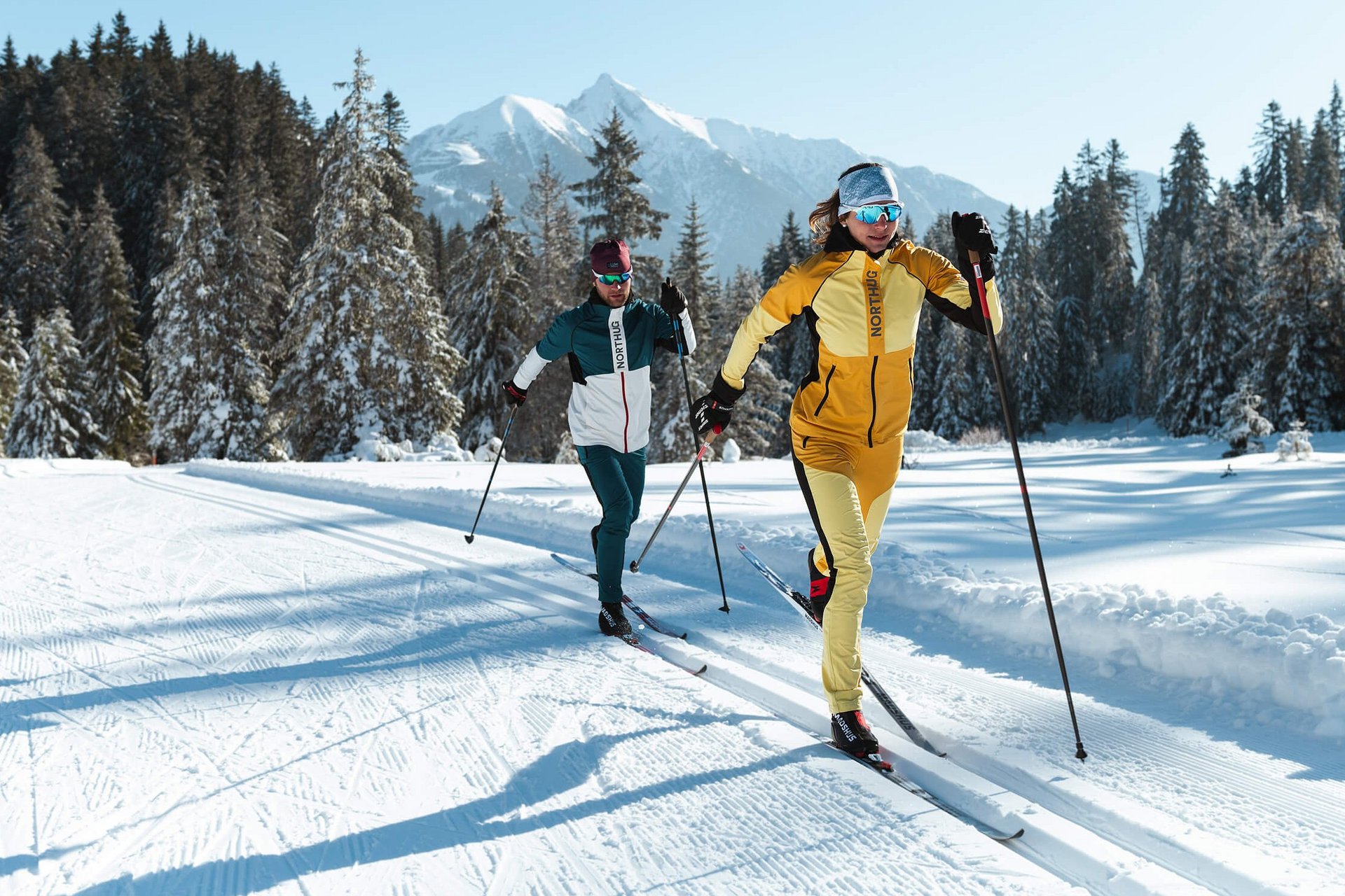 Zwei Langläufer in bunter Ausrüstung auf verschneiter Loipe vor Bergen