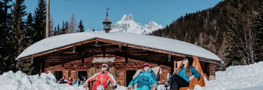 Pause auf der Skitour in Filzmoos © Nadia Jabli_www.filzmoos.at Drei Personen entspannen sich im Schnee vor einer Berghütte unter blauem Himmel