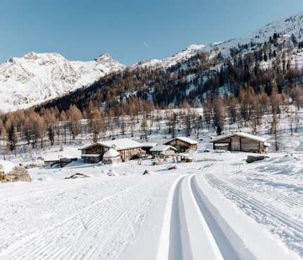 Langlaufloipe führt zu verschneiten Hütten und Bergen unter klarem Himmel