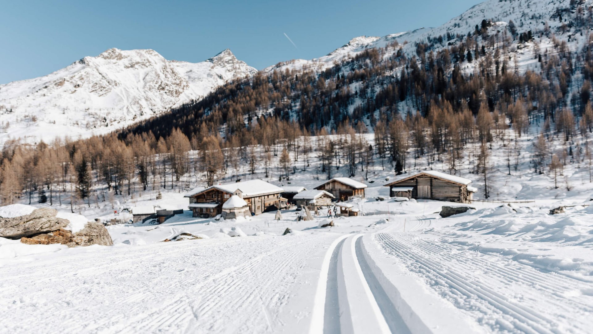 Langlaufloipe führt zu verschneiten Hütten und Bergen unter klarem Himmel