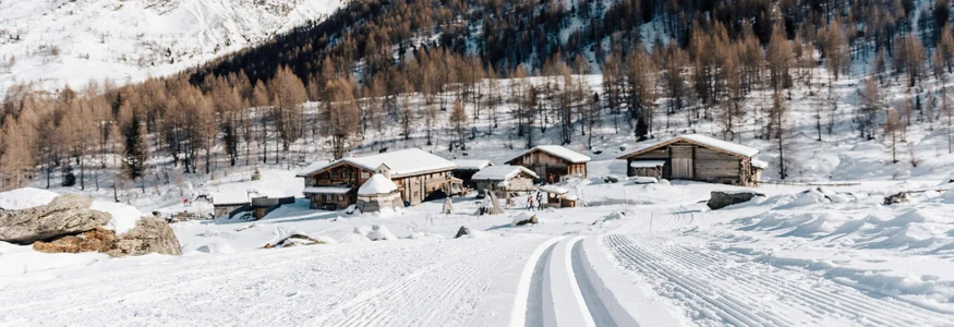 Langlaufloipe führt zu verschneiten Hütten und Bergen unter klarem Himmel