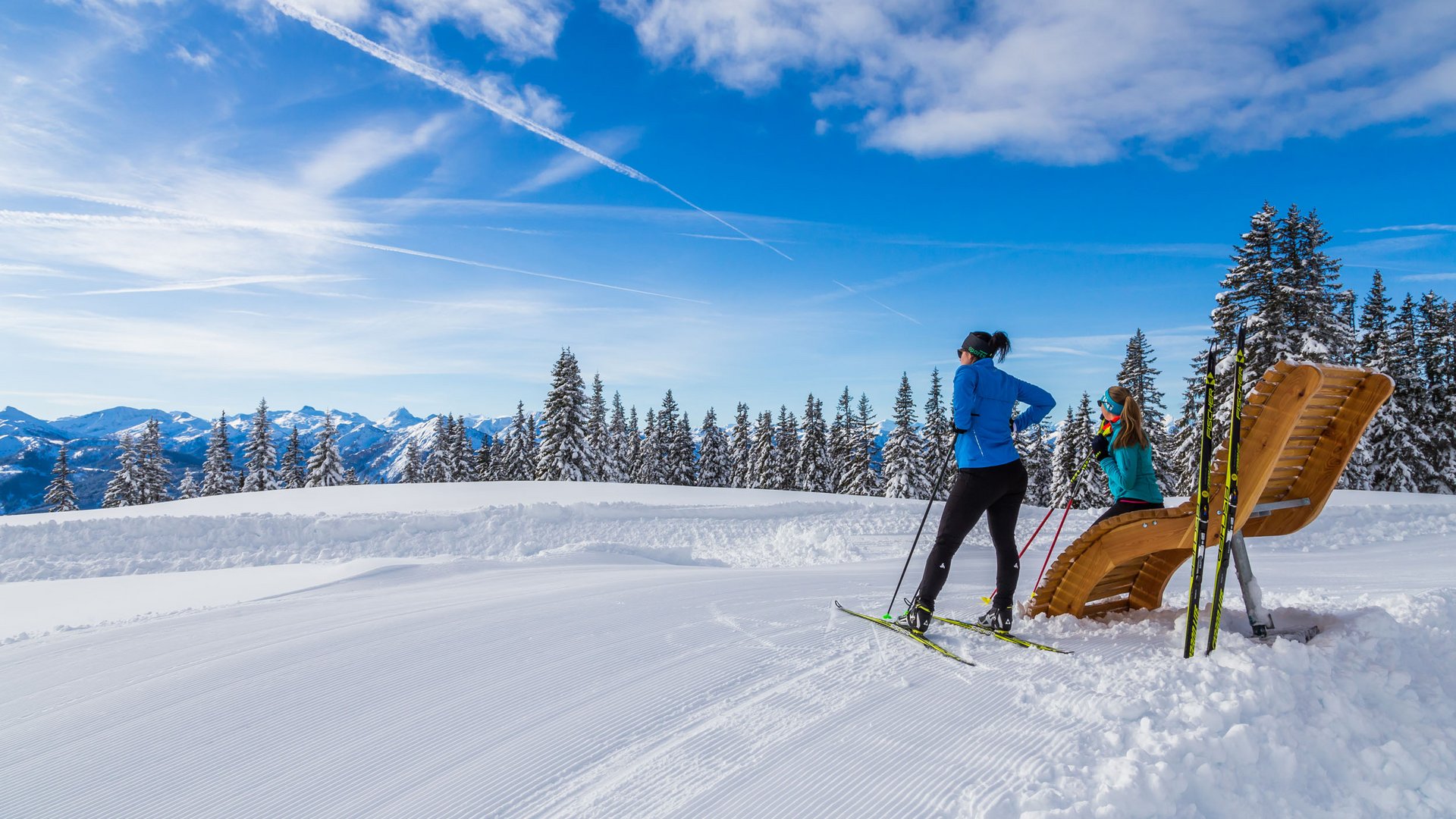 Two cross-country skiers in snow with snowy trees and mountains in background