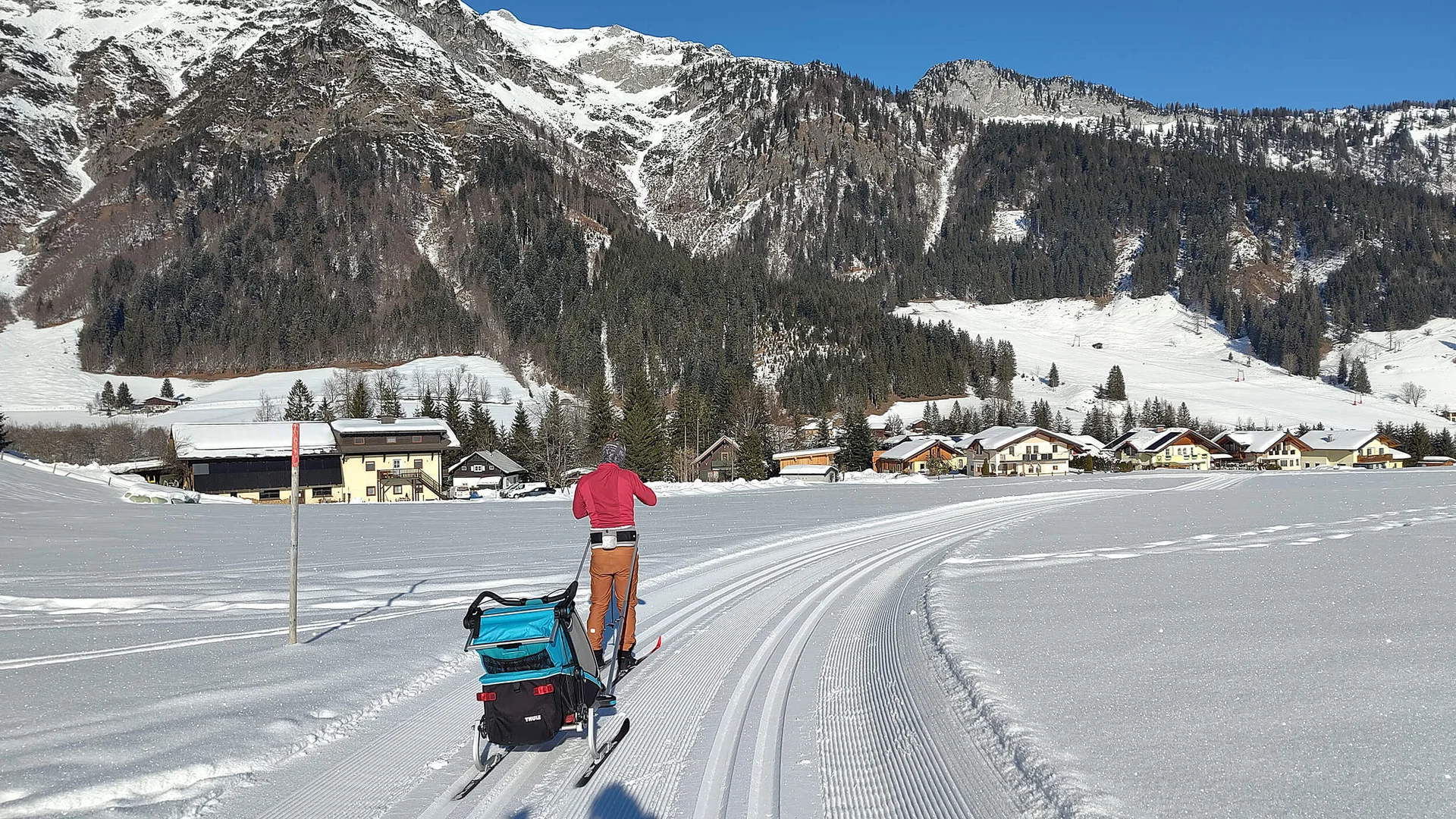 Langläufer mit Kinderwagen unterwegs auf präparierter Loipe vor Bergkulisse
