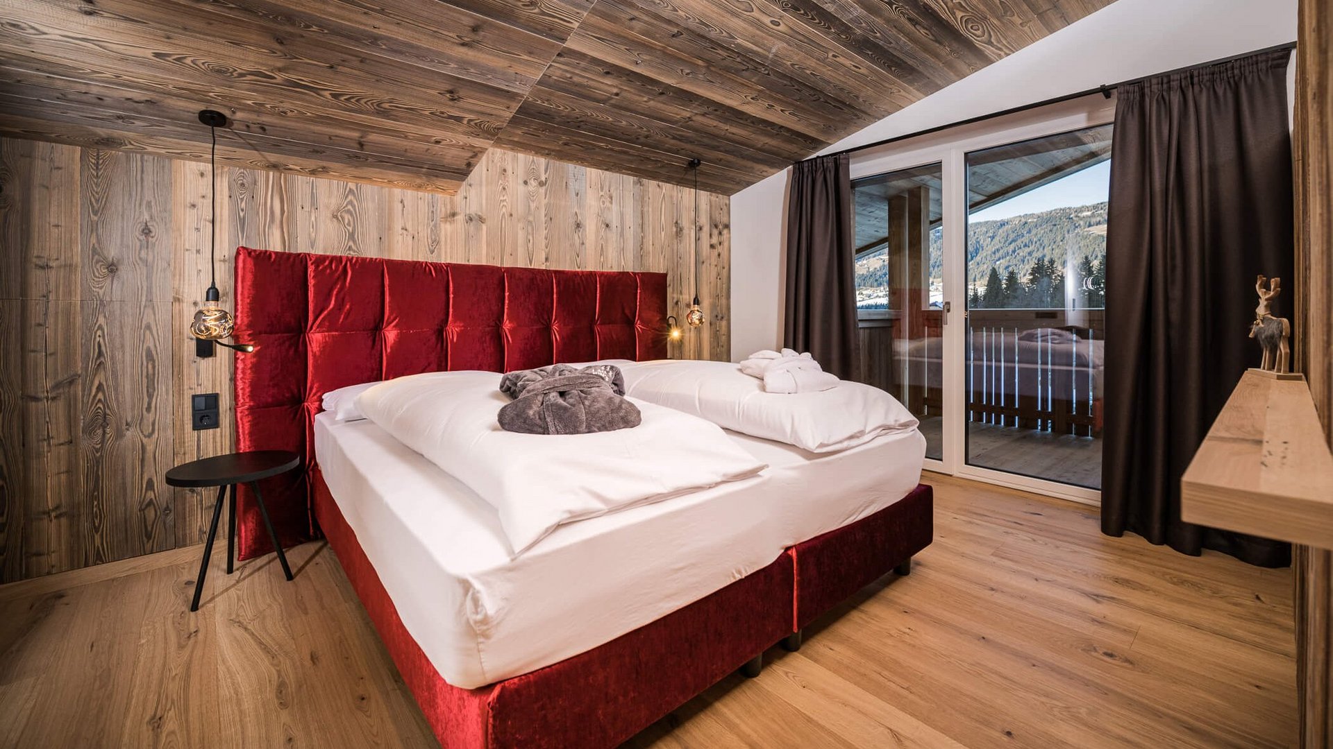 Schlafzimmer mit rotem Bett und Holzdecke, Blick auf Balkon und Berge