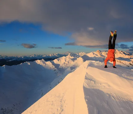 Person macht Handstand auf verschneitem Berggipfel bei Sonnenuntergang
