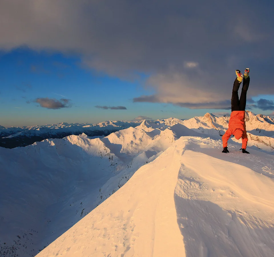 Person macht Handstand auf verschneitem Berggipfel bei Sonnenuntergang