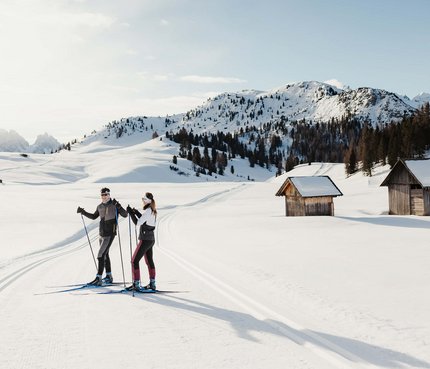 Zwei Skifahrer auf gespurter Loipe in verschneiter Berglandschaft mit Hütten