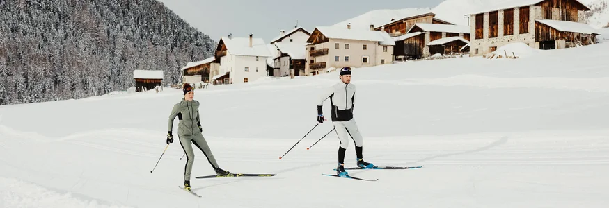 Zwei Skifahrer beim Langlauf vor verschneiten Häusern und Wald