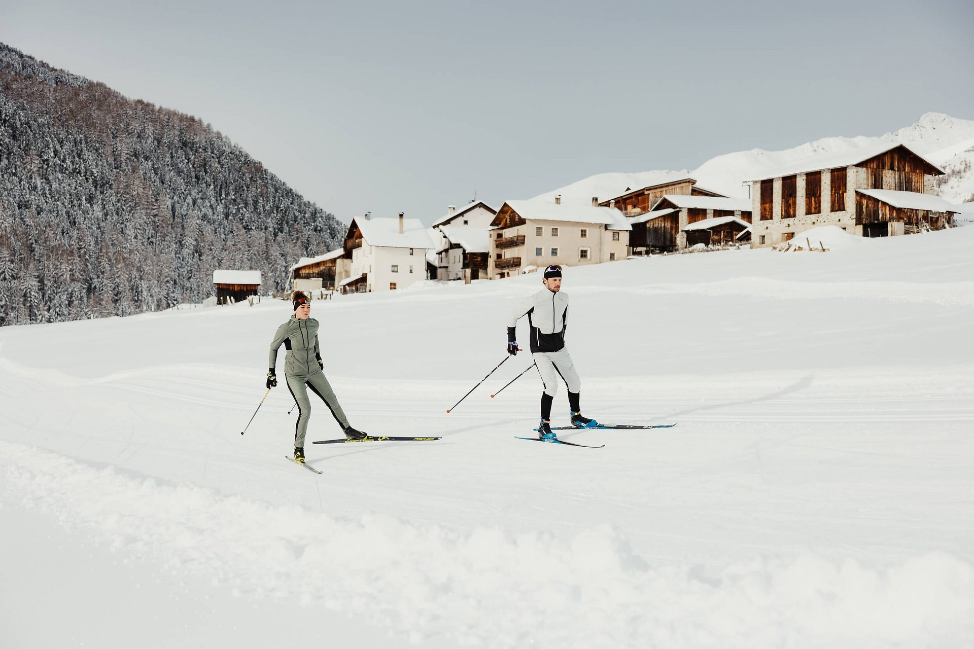 Zwei Skifahrer beim Langlauf vor verschneiten Häusern und Wald