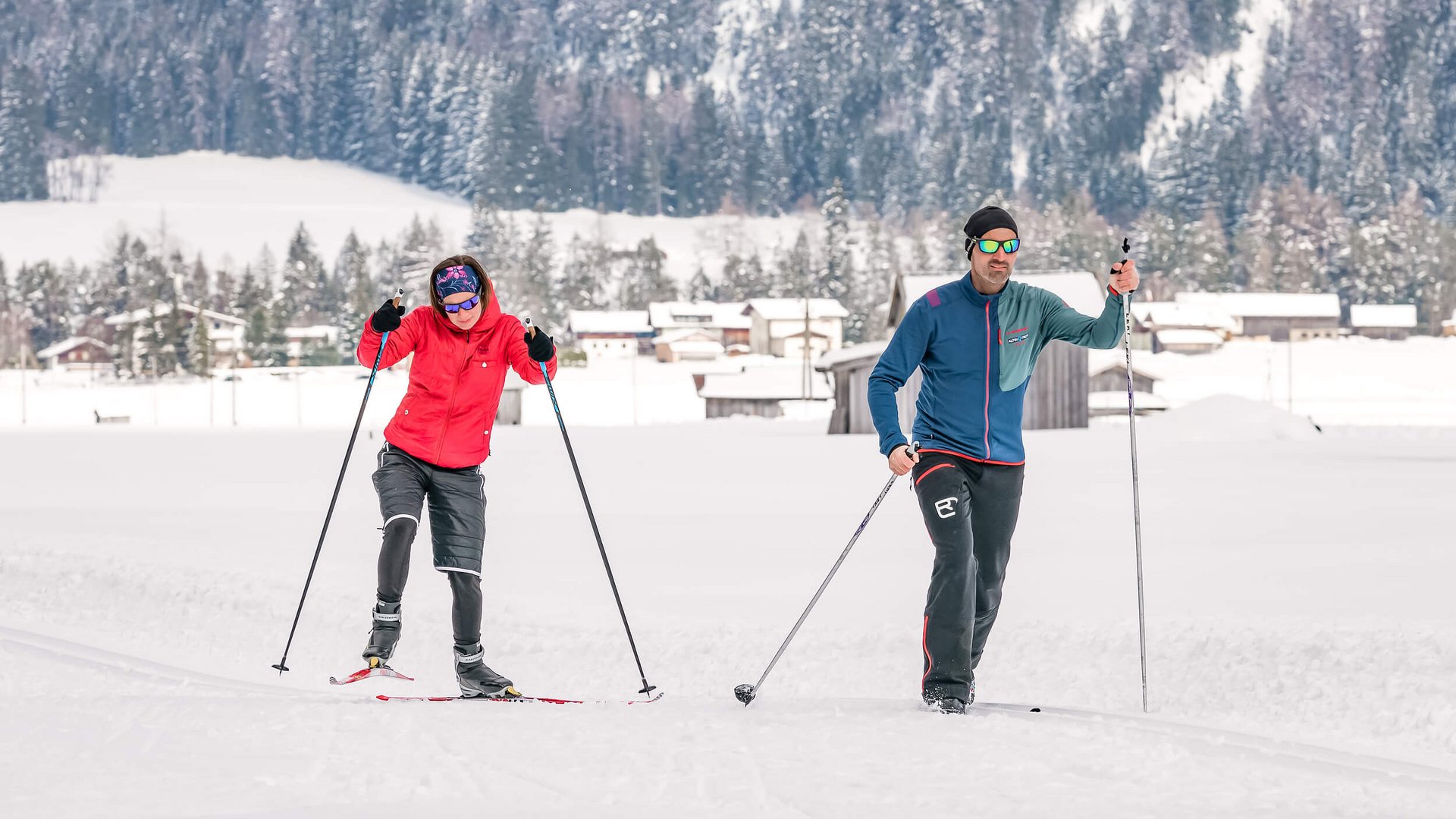 Zwei Personen beim Langlaufen auf verschneiter Landschaft mit Häusern und Wäldern im Hintergrund