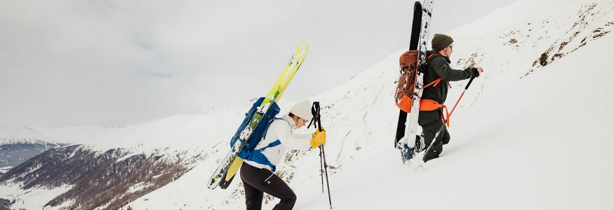 Zwei Skifahrer steigen mit Skiern am Rücken einen schneebedeckten Hang hinauf.