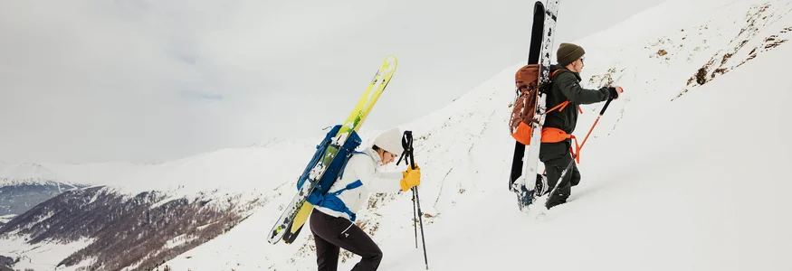 Zwei Skifahrer steigen mit Skiern am Rücken einen schneebedeckten Hang hinauf.