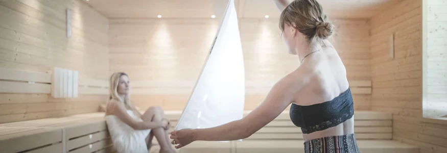 Zwei Frauen in einem hellen Holzsauna, eine sitzt, die andere hält ein Handtuch hoch