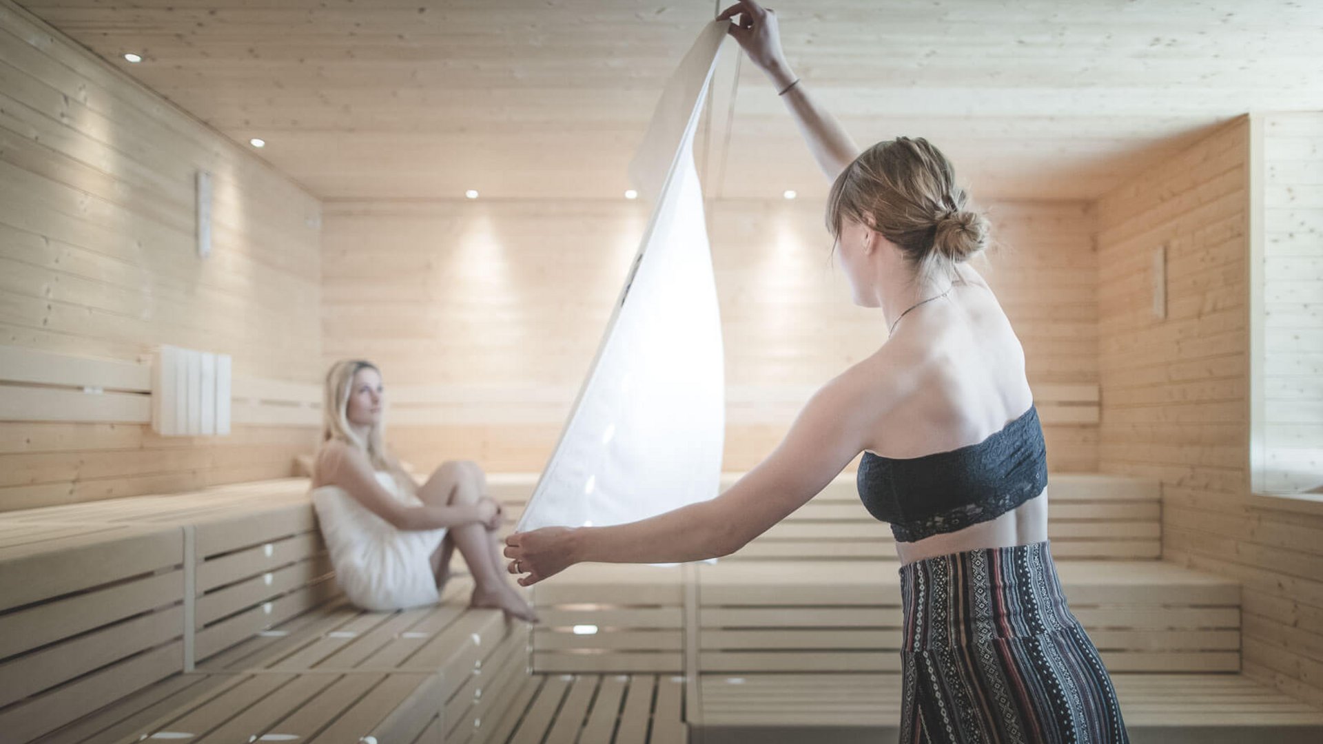 Zwei Frauen in einem hellen Holzsauna, eine sitzt, die andere hält ein Handtuch hoch