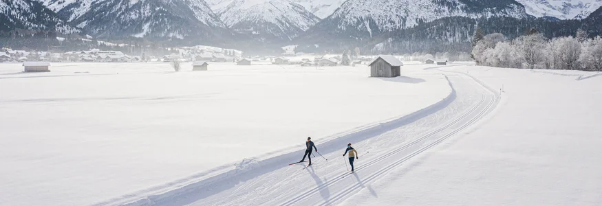 Zwei Skifahrer auf einer Loipe vor verschneiten Bergen bei Sonnenschein