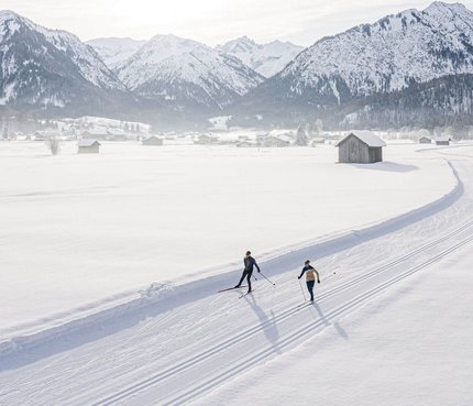 Zwei Skifahrer auf einer Loipe vor verschneiten Bergen bei Sonnenschein