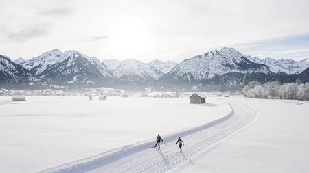 Two skiers on a trail in a snowy landscape with mountains and sun