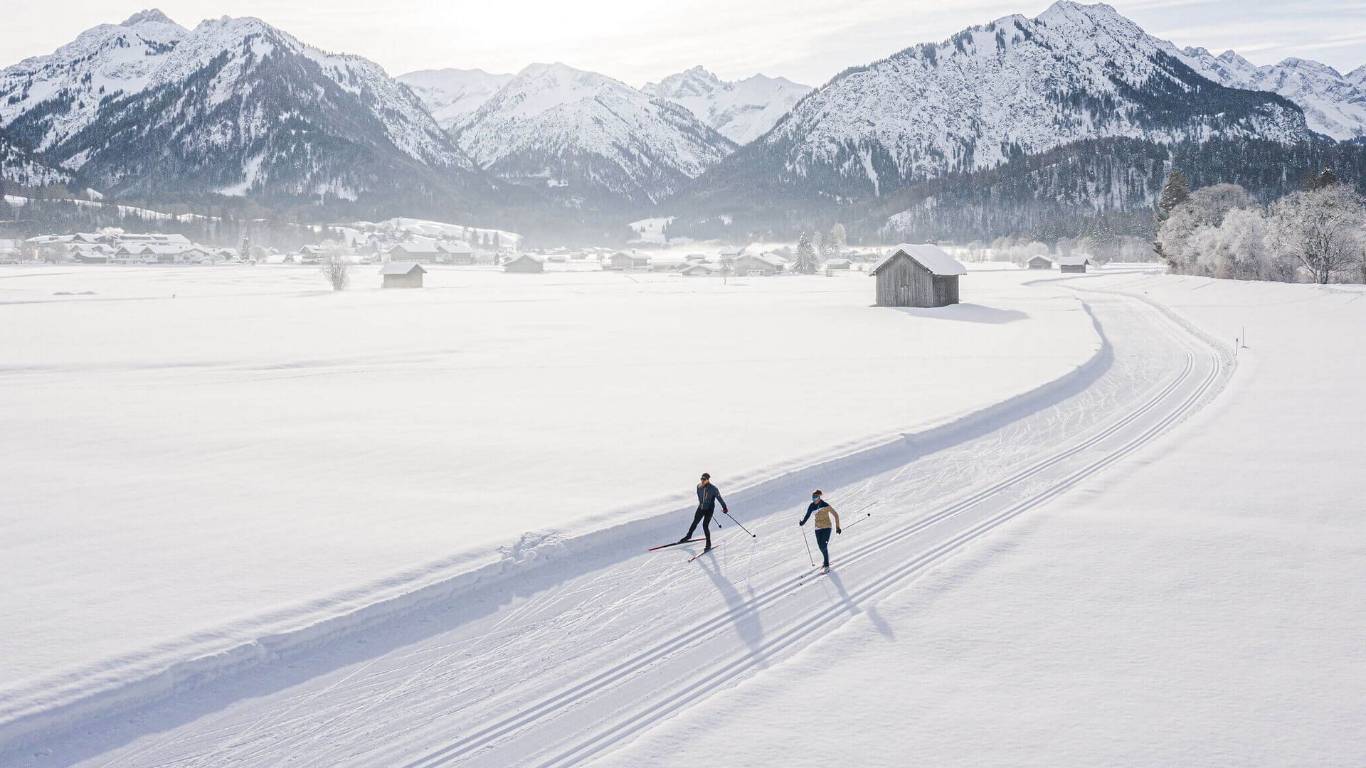 Two skiers on a trail in a snowy landscape with mountains and sun