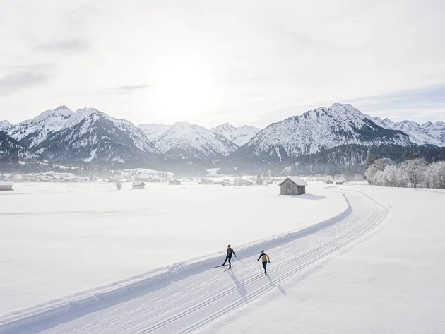 Two skiers on a trail in a snowy landscape with mountains and sun