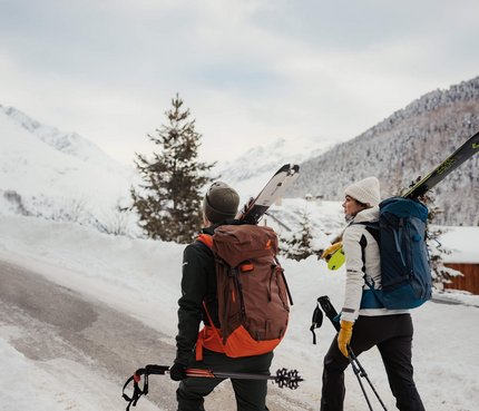 Zwei Skifahrer mit Rucksäcken und Skiern gehen einen schneebedeckten Bergweg hinauf