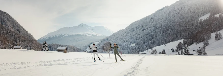 nordic³ © Alex Moling Zwei Langläufer auf verschneiter Strecke vor bewaldeten Bergen bei Sonnenlicht
