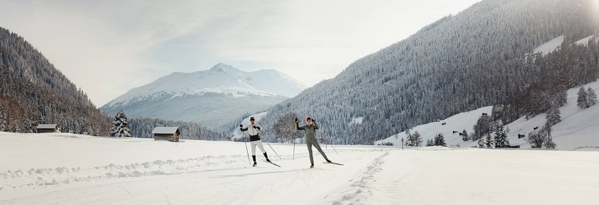 Zwei Langläufer auf verschneiter Strecke vor bewaldeten Bergen bei Sonnenlicht