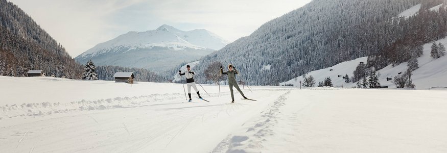 Zwei Langläufer auf verschneiter Strecke vor bewaldeten Bergen bei Sonnenlicht
