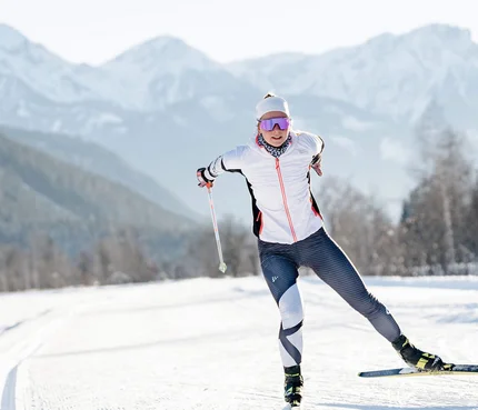 Langläuferin in weißer Jacke auf verschneiter Loipe vor Bergen