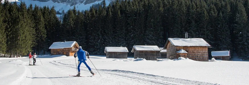 Cross-country skiers near snowy cabins with mountain backdrop on sunny winter day