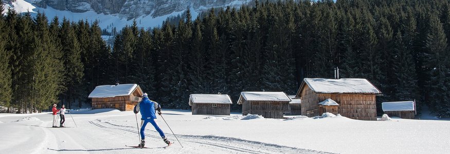 Cross-country skiers near snowy cabins with mountain backdrop on sunny winter day