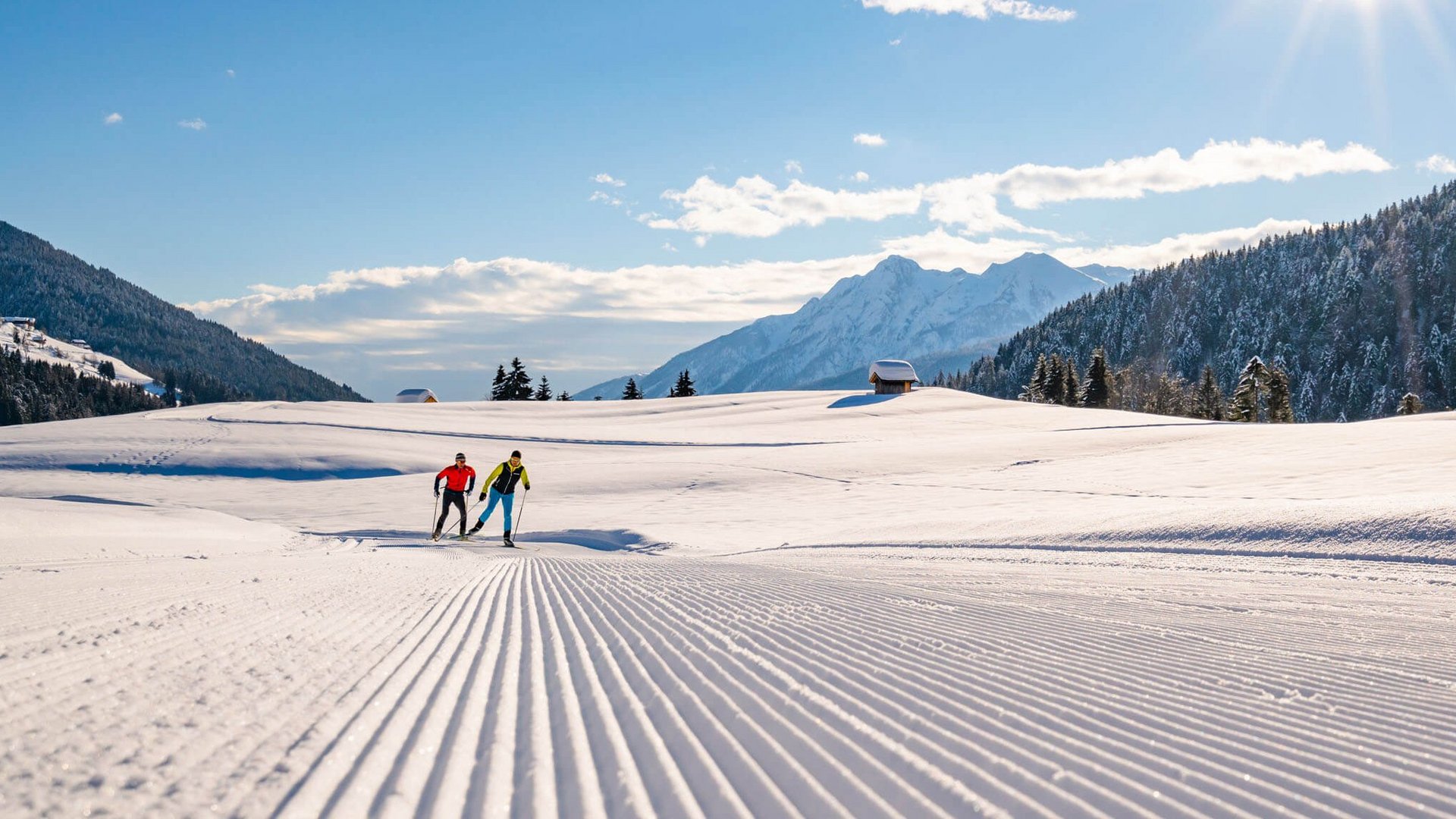 Two cross-country skiers on groomed snow trail in sunny mountain landscape
