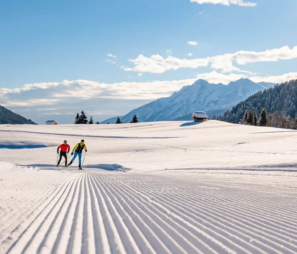 Two cross-country skiers on groomed snow trail in sunny mountain landscape
