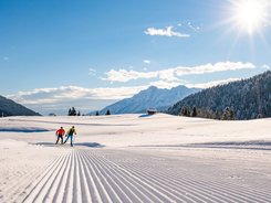 Two cross-country skiers on groomed snow trail in sunny mountain landscape