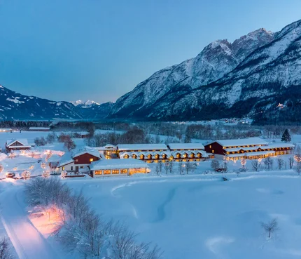 Snow-covered houses in valley with lit mountains at dusk