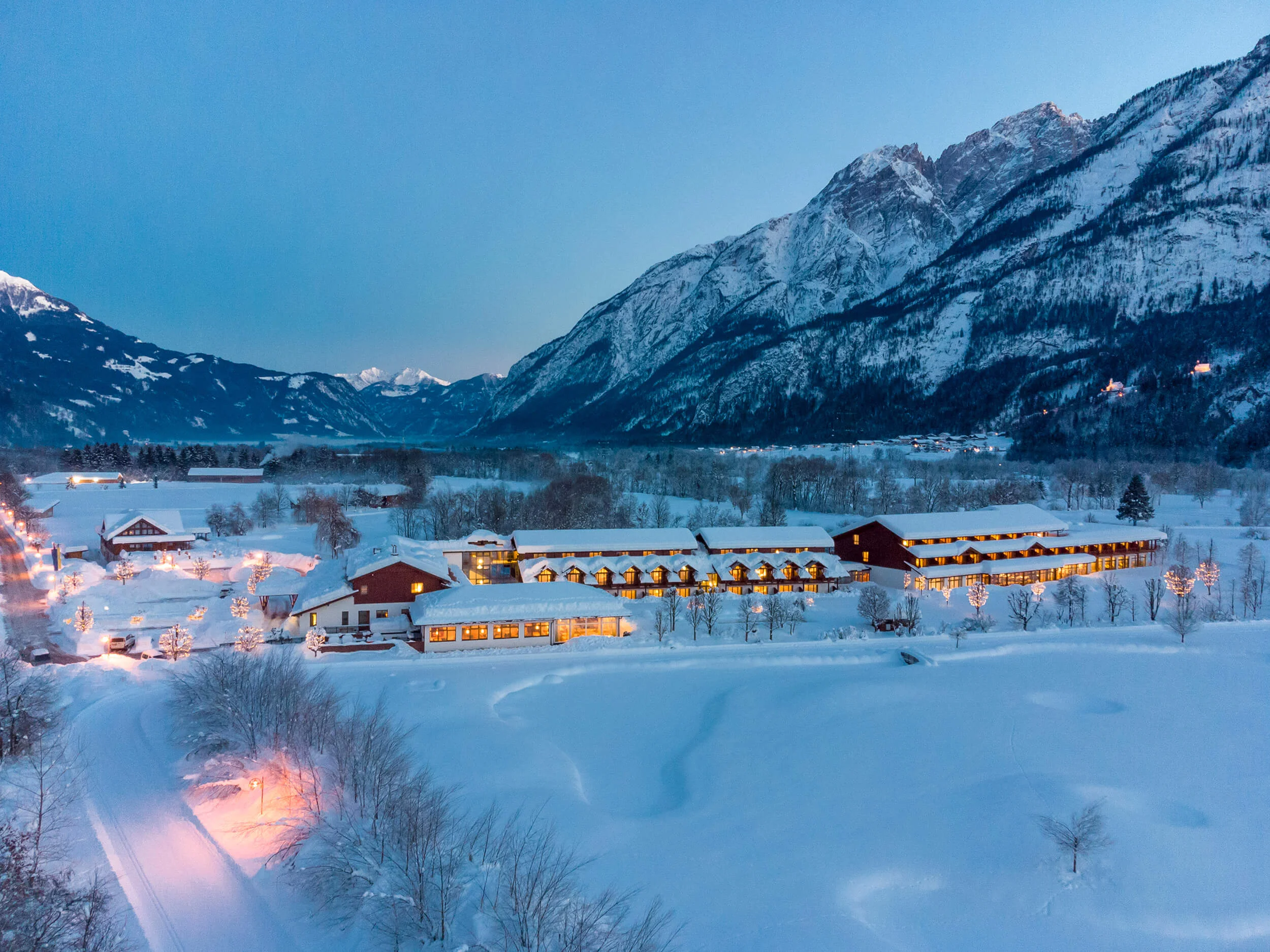 Snow-covered houses in valley with lit mountains at dusk