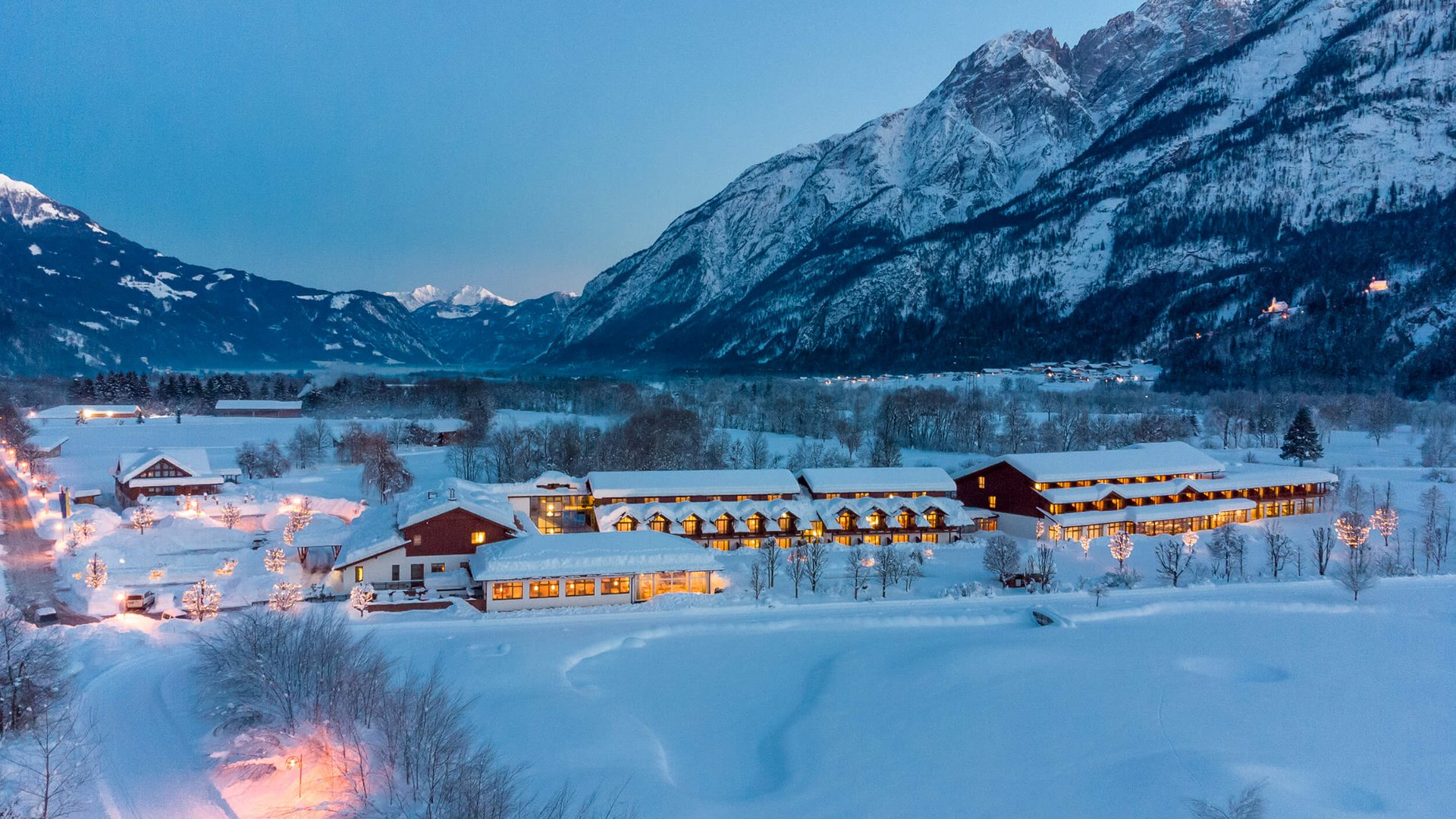 Snow-covered houses in valley with lit mountains at dusk