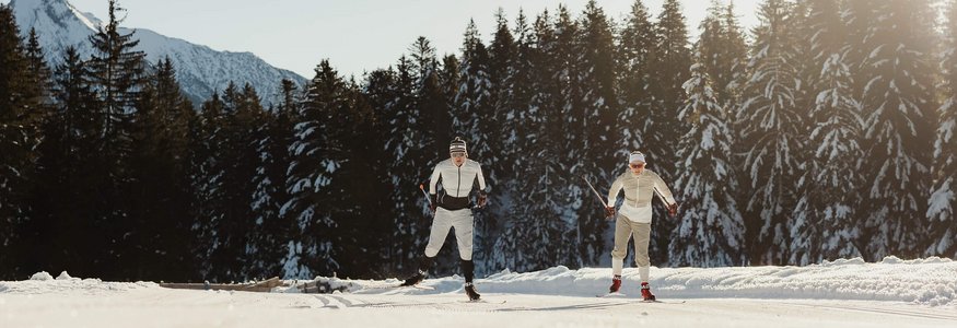 Two cross-country skiers on snowy trail with forest and mountains