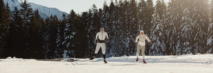 Zwei Skilangläufer auf verschneiter Strecke vor Wald und Bergkulisse