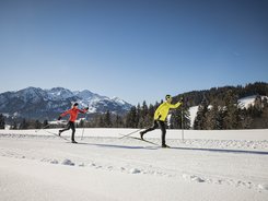 Two people cross-country skiing on snow with mountains in background