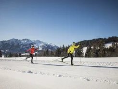 Two people cross-country skiing on snow with mountains in background