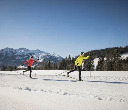 Two people cross-country skiing on snow with mountains in background