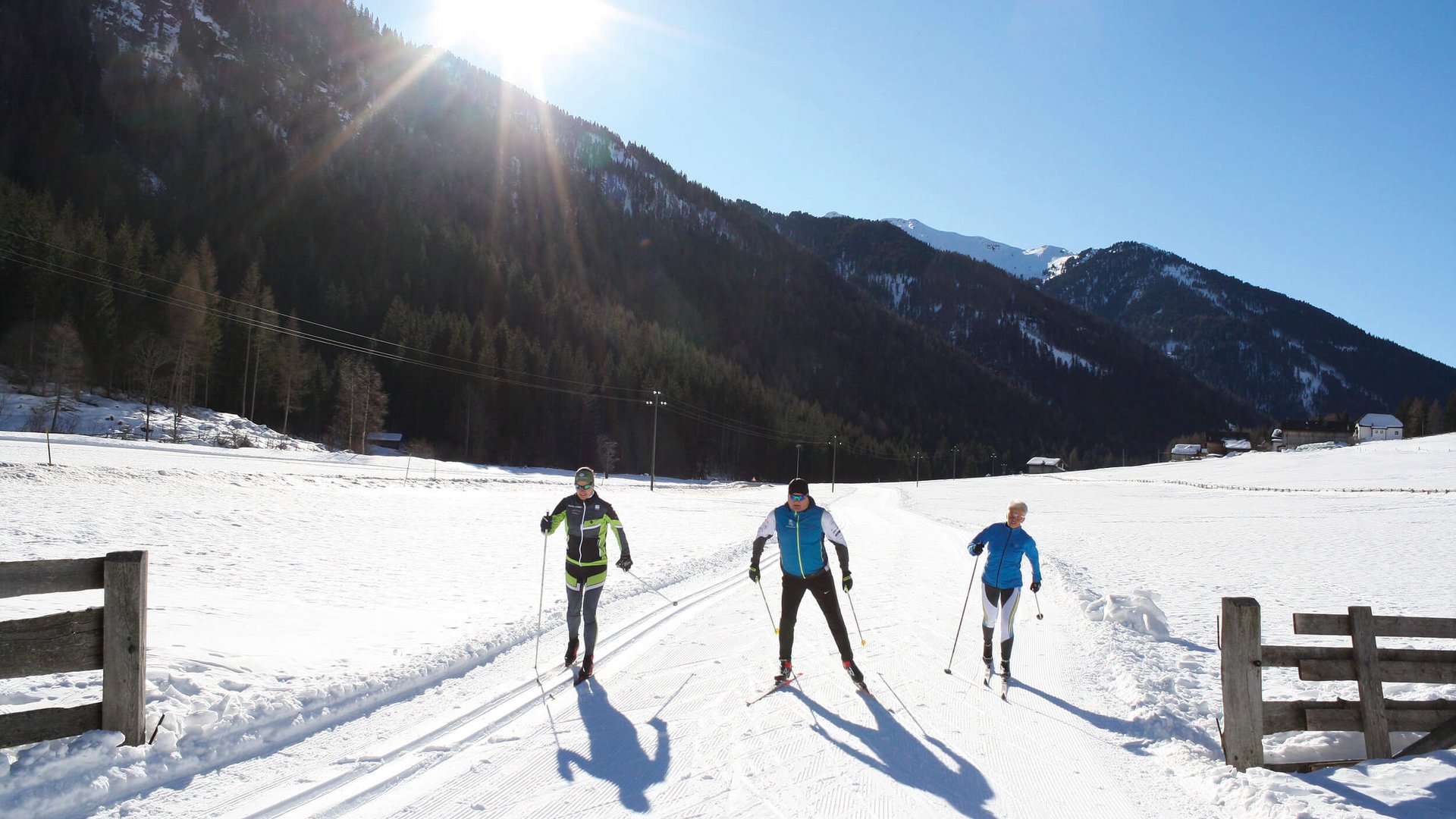 Drei Langläufer auf schneebedeckter Strecke mit Bergen und Sonne im Hintergrund