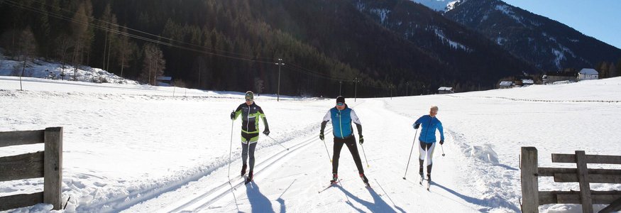 Drei Langläufer auf schneebedeckter Strecke mit Bergen und Sonne im Hintergrund
