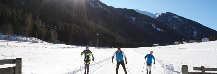Drei Langläufer auf schneebedeckter Strecke mit Bergen und Sonne im Hintergrund