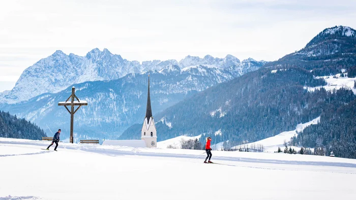 Zwei Langläufer in verschneiter Landschaft mit Kirche und Bergen im Hintergrund
