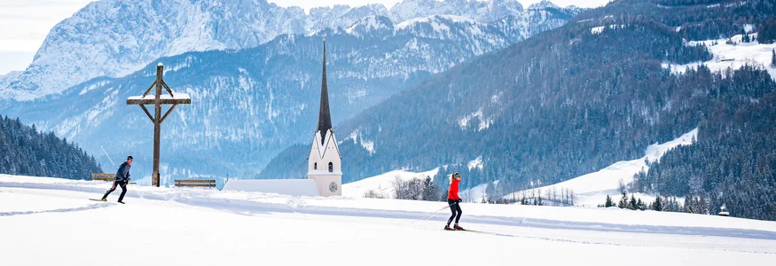 Zwei Langläufer in verschneiter Landschaft mit Kirche und Bergen im Hintergrund