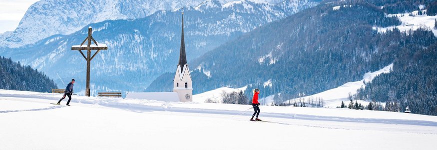 Zwei Langläufer in verschneiter Landschaft mit Kirche und Bergen im Hintergrund